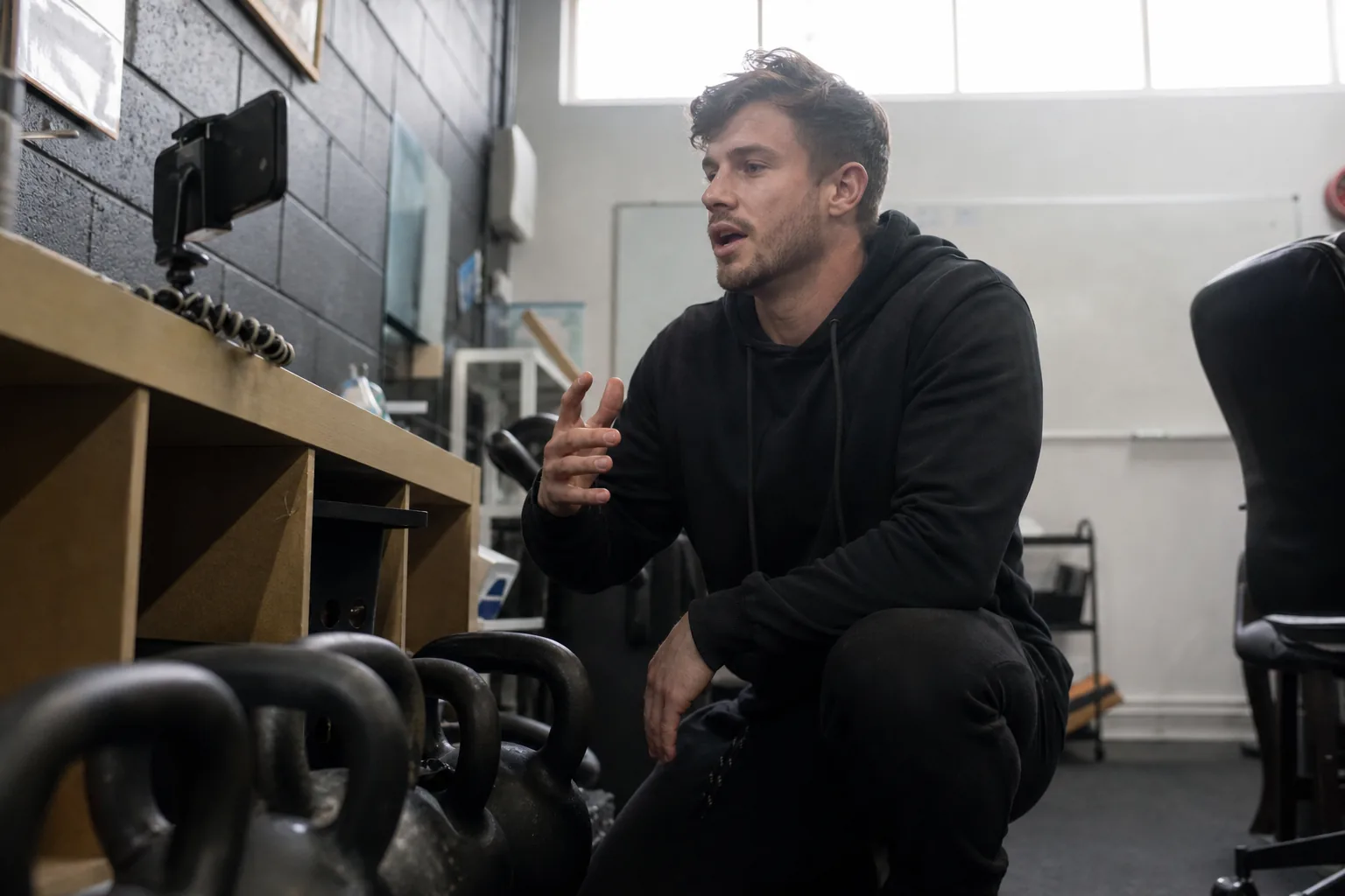 Fitness coach filming a short social clip on a phone clamped to a shelf in a small gym office