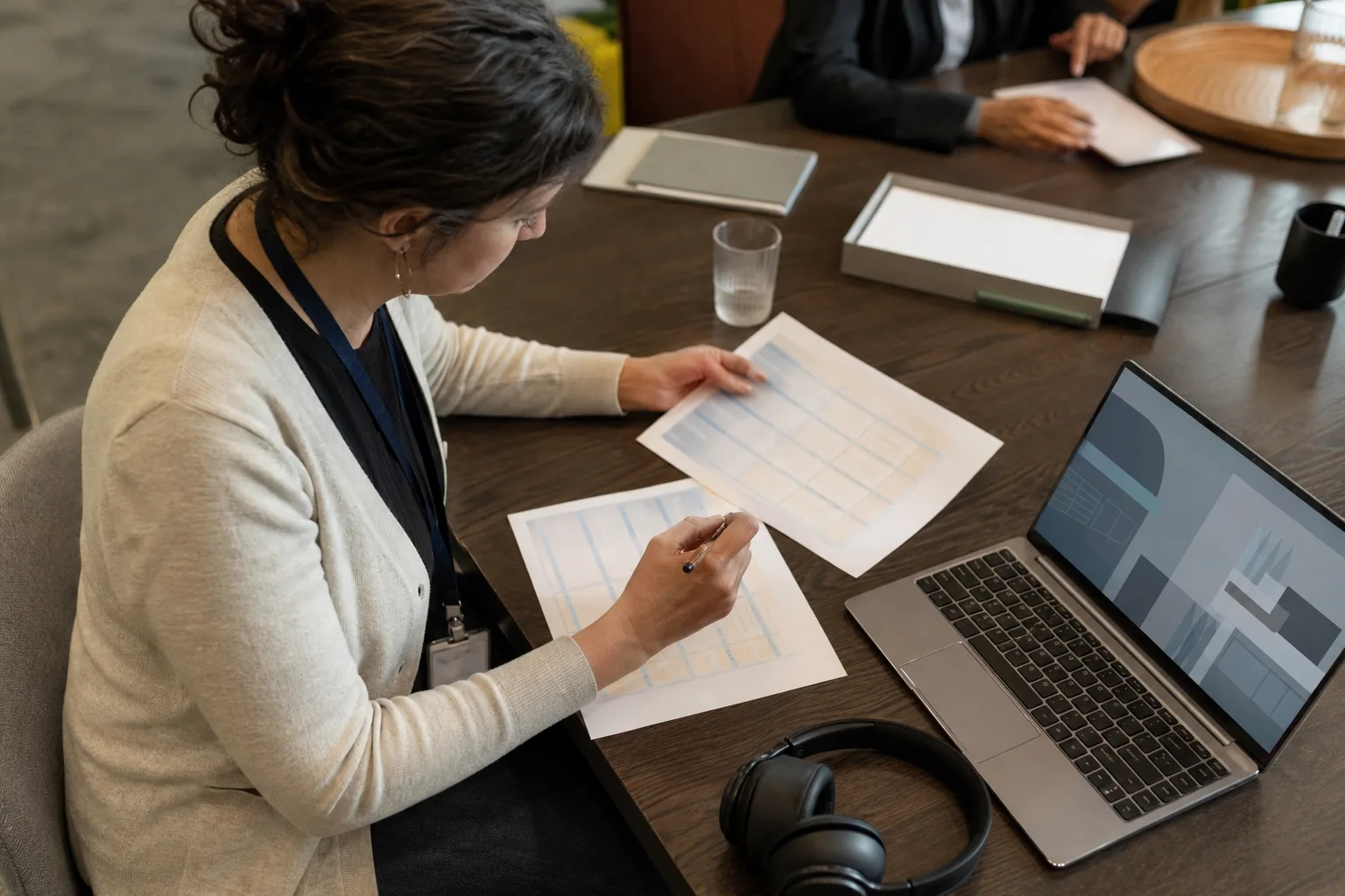 Compliance officer comparing printed vendor checklists with headphones and a laptop on the table