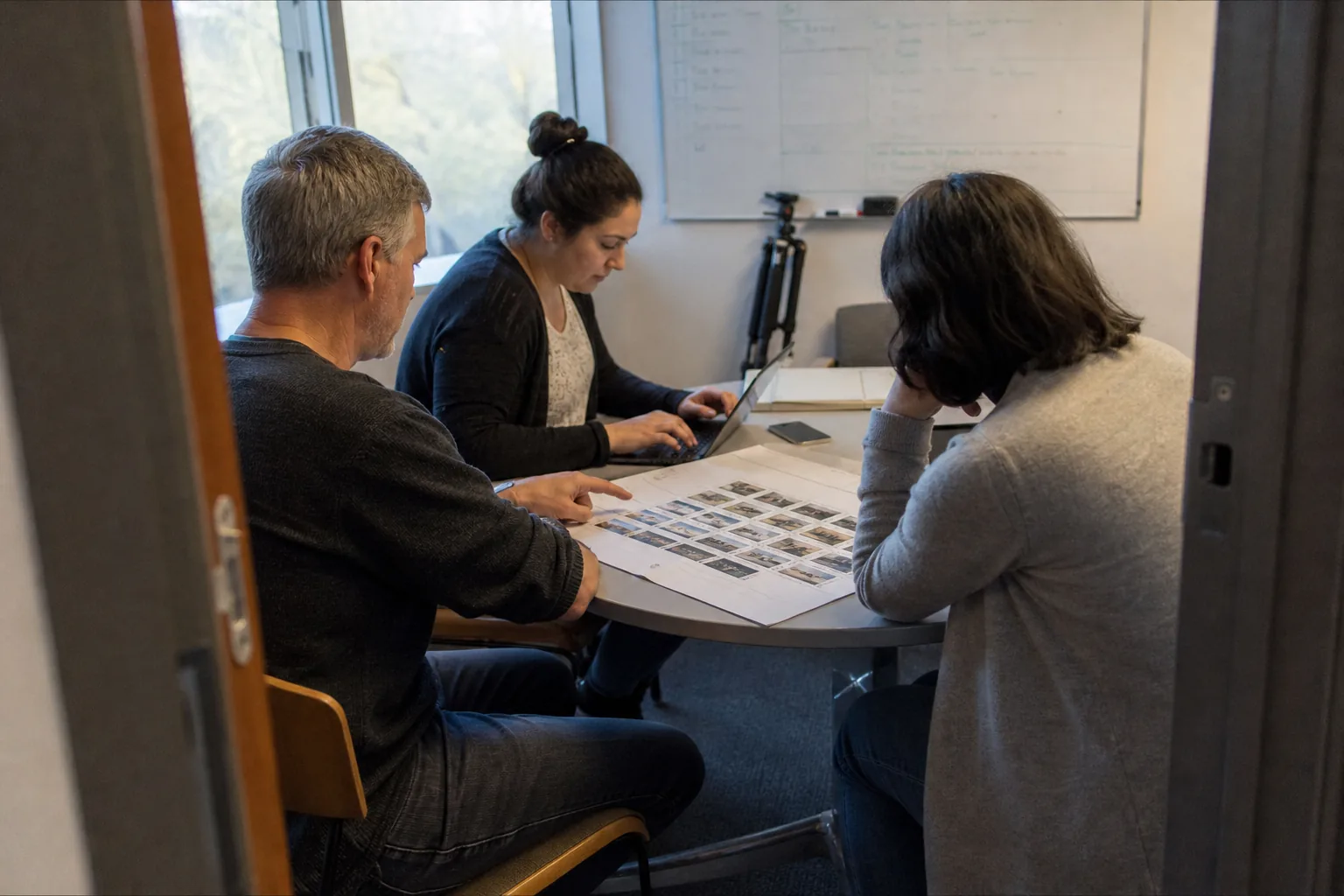 Learning and development team around a table reviewing a printed storyboard and a laptop with a script, a folded tripod in the corner